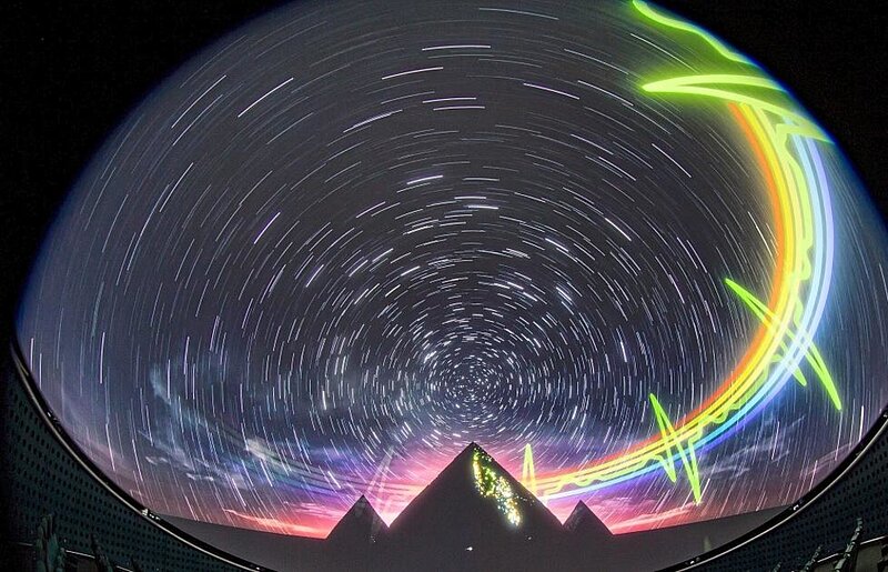 Kuppelbild einer Langzeitaufnahme des Sternenhimmels mit Stern-Strichspuren hinter scherenschnittartigen Pyramiden mit pulsierenden regenbogenfarbenen Streifen