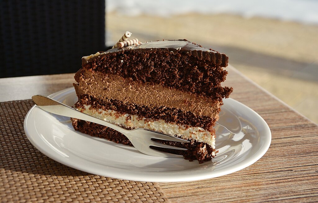 Ein großes Stück Nougattorte auf einem Holztisch angerichtet auf einem weißen Teller mit Kuchengabel.