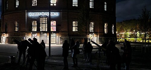 Menschen mit mobilen Teleskopen im Schattenriss beobachten den Nachthimmel vor dem nächtlich beleuchteten Planetarium