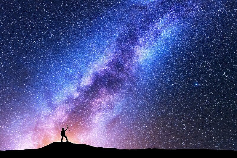 Sternenübersäter blauer Nachthimmel mit blau-weiß-rötliche schimmernder Milchstraße, davor scherenschnittartige Höhenlandschaft mit einer zum Himmel aufschauenden Person.