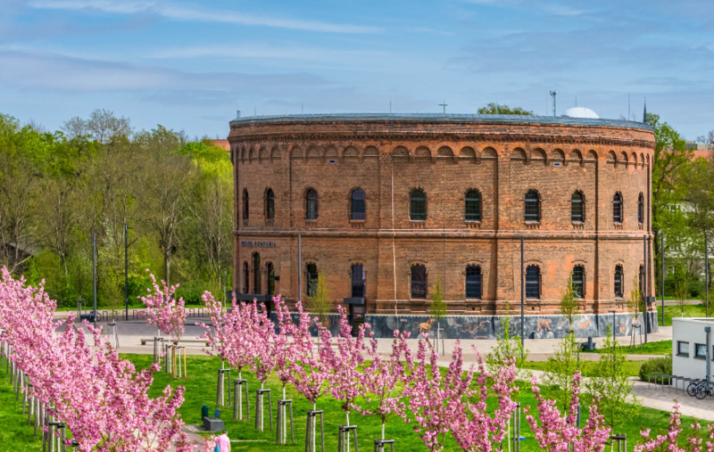 Historisches Gasometer-Rundgebäude mit Ziegelfassade und zahlreichen Fenstern hinter einer Allee rosafarbener Blütenbäumen