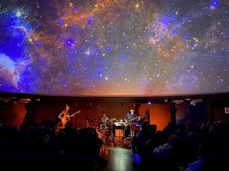 2 Gitarristen und Schlagzeuger in Planetarium mit blau-roter Kuppelprojektion