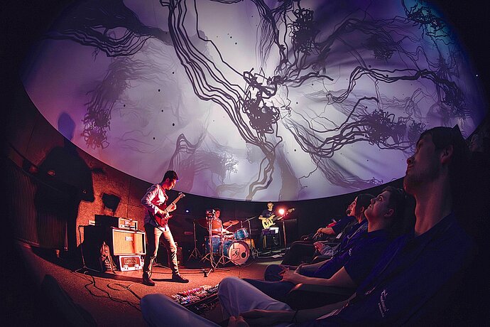 Band mit Gitarristen und Schlagzeug vor Zuschauern im Planetarium mit Projektion in der Kuppel