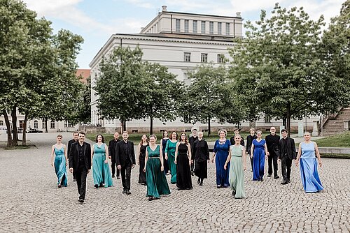 Chorsänger in schwarzer, grüner und blauer festlicher Kleidung vor Universitätsgebäude unter grünen Bäumen