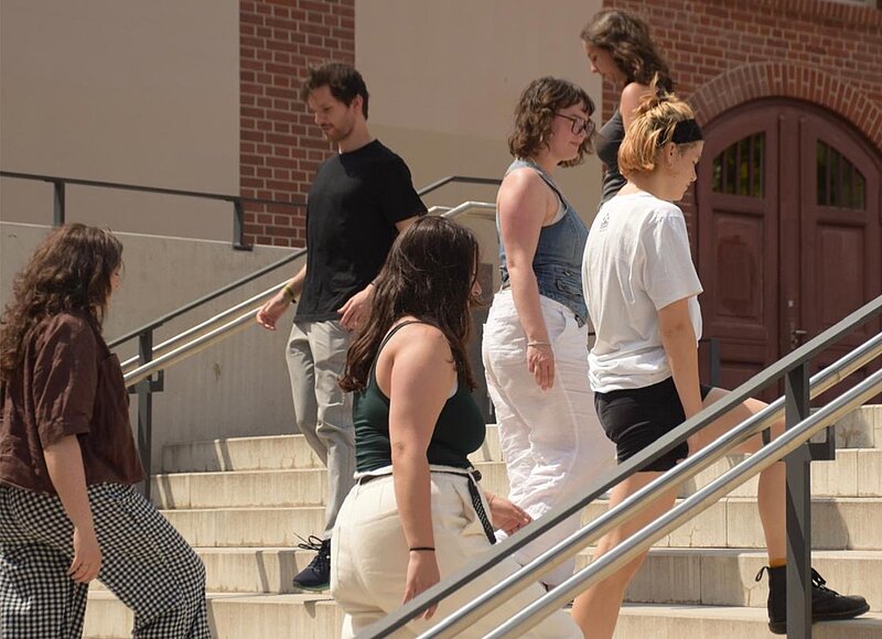 Junge Menschen, die im Rahmen einer szenischen Darstellung vor einem Universitätsgebäude auf einer Treppe auf- und abgehen.