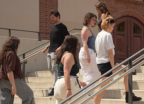 Junge Menschen, die im Rahmen einer szenischen Darstellung vor einem Universitätsgebäude auf einer Treppe auf- und abgehen.