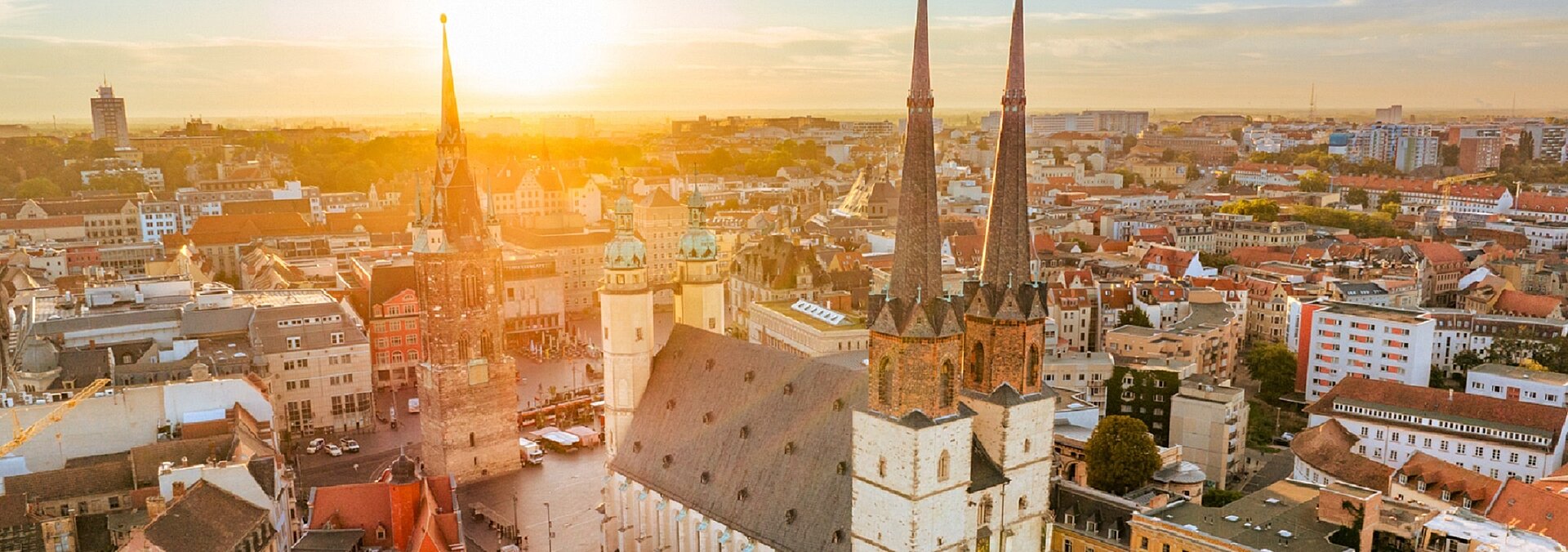 Stimmungsvolles Luftbild vom Halleschen Marktplatz im Gegenlicht mit Marktkirche und durch die tiefstehendes Sonne orange angestrahlten Häusern FuenfTuerme_Markt_Platz2023_SMG_Header.jpg