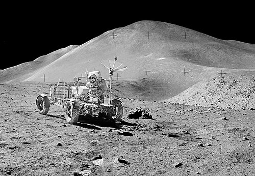 schwarz-weiß Foto mit Astronaut an vierrädrigem Mondfahrzeug vor einem Hügel auf dem Erdmond