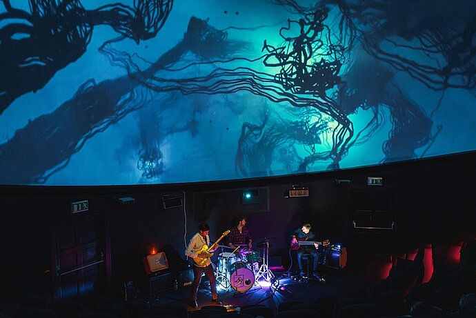 2 Gitarristen und Schlagzeuger vor Kuppelprojektion