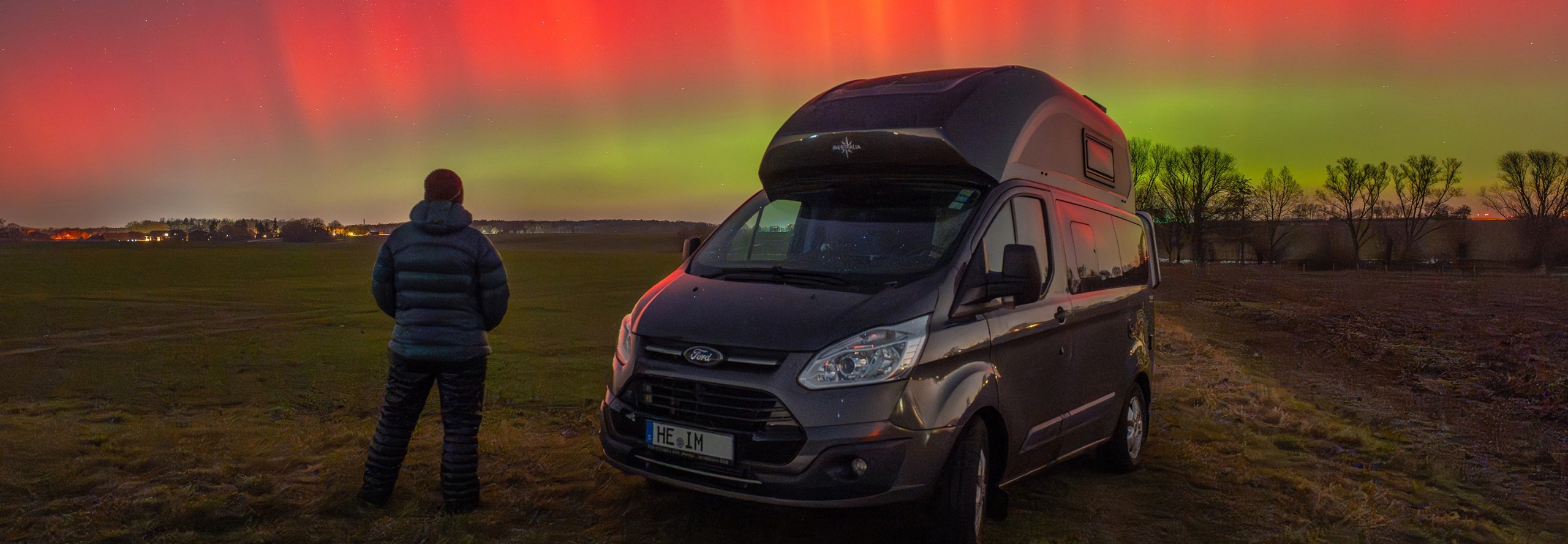 Eine Person steht mit einem Camper auf einem dunklen Feld und im Hintergrund sind am Himmel Polarlichter zu sehen.
