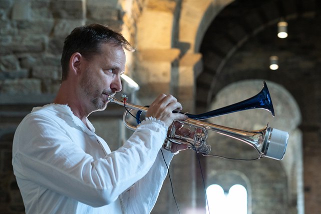 Portrait eines Mannes mit leichtem Vollbart, kurzem Haar, bekleidet mit weißem langärmeligen Oberteil mit Trompete in einem beleuchteten Kreuzgang