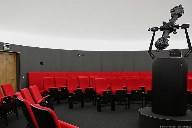 Im Sternsaal des Planetariums stehen Planetariumssessel mit rotem Polster vor dunkler Wand. Rechts im Vordergrund der Sternprojektor, der sich vor der weißen Sternkuppel kontrastreich abhebt.