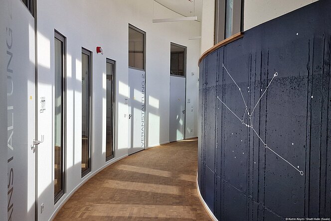 Flur im Obergeschoss des Planetariums mit Parkettboden, blaue Wand mit weißer Sternbild-Darstellung des Orions. Linkerhand Wand, Türen und Sichtfenster zu Veranstaltungsräumen mit Licht- und Schattenspiel.